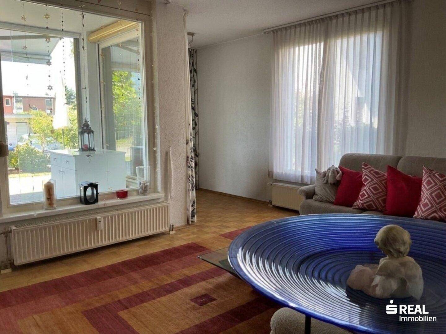 Helles Wohnzimmer mit großen Fenstern, Teppichboden und einem beigen Sofa.