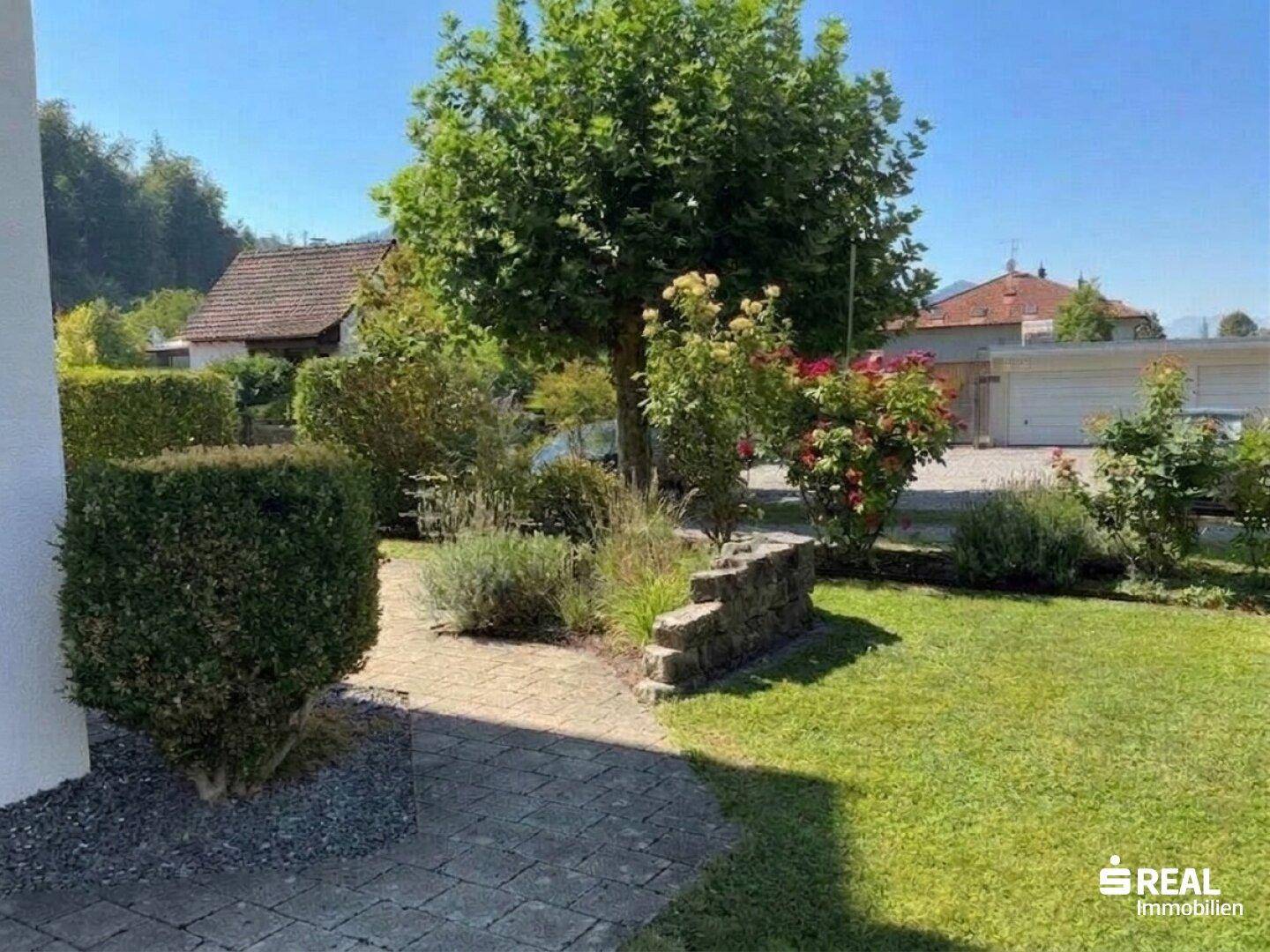 Gepflegter Gartenbereich mit Rasenfläche, Blumenbeeten und einem großen Baum im Vordergrund.