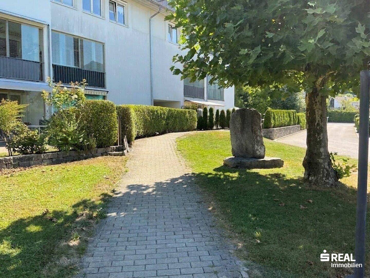 Gepflasterter Weg entlang des Gebäudes, umgeben von Rasenflächen und Hecken.