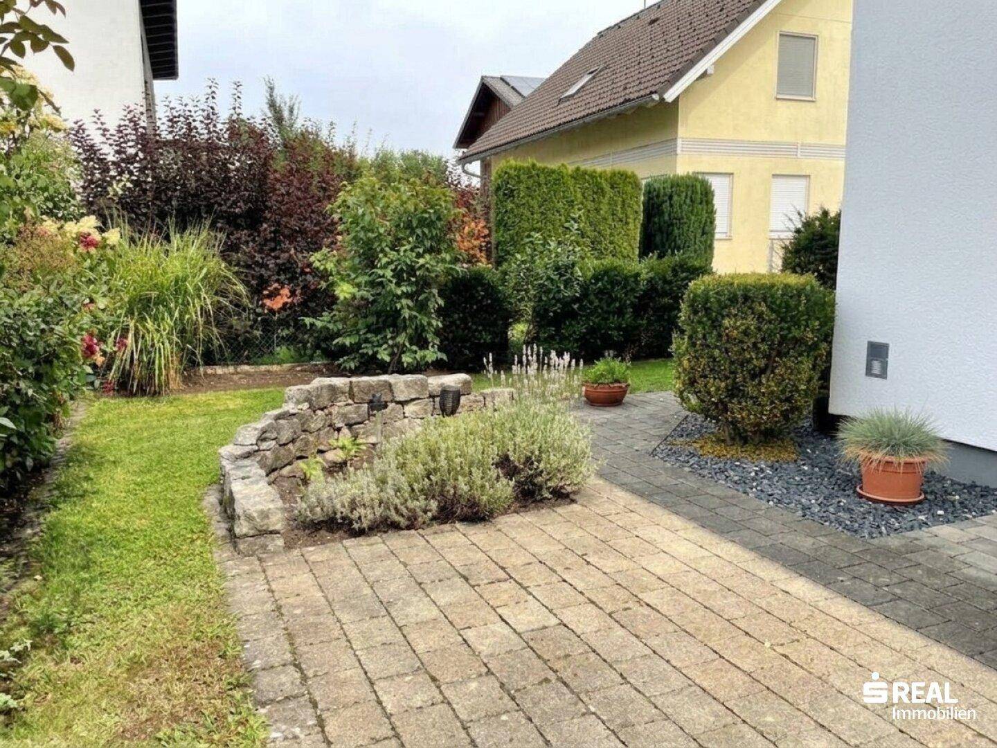 Seitlicher Gartenbereich mit gepflastertem Weg, Rasen und einer Steinmauer als Beeteinfassung.