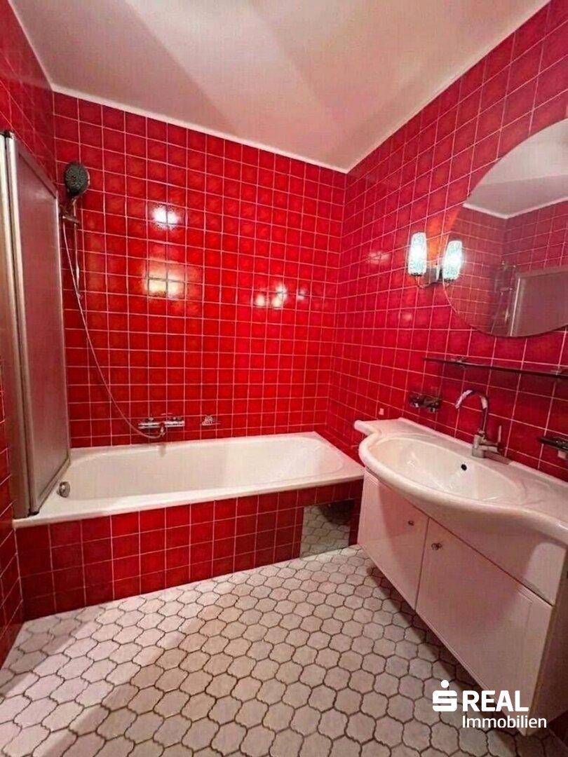 Badezimmer mit roten Wandfliesen, Badewanne mit Duschabtrennung und Einzelwaschbecken.