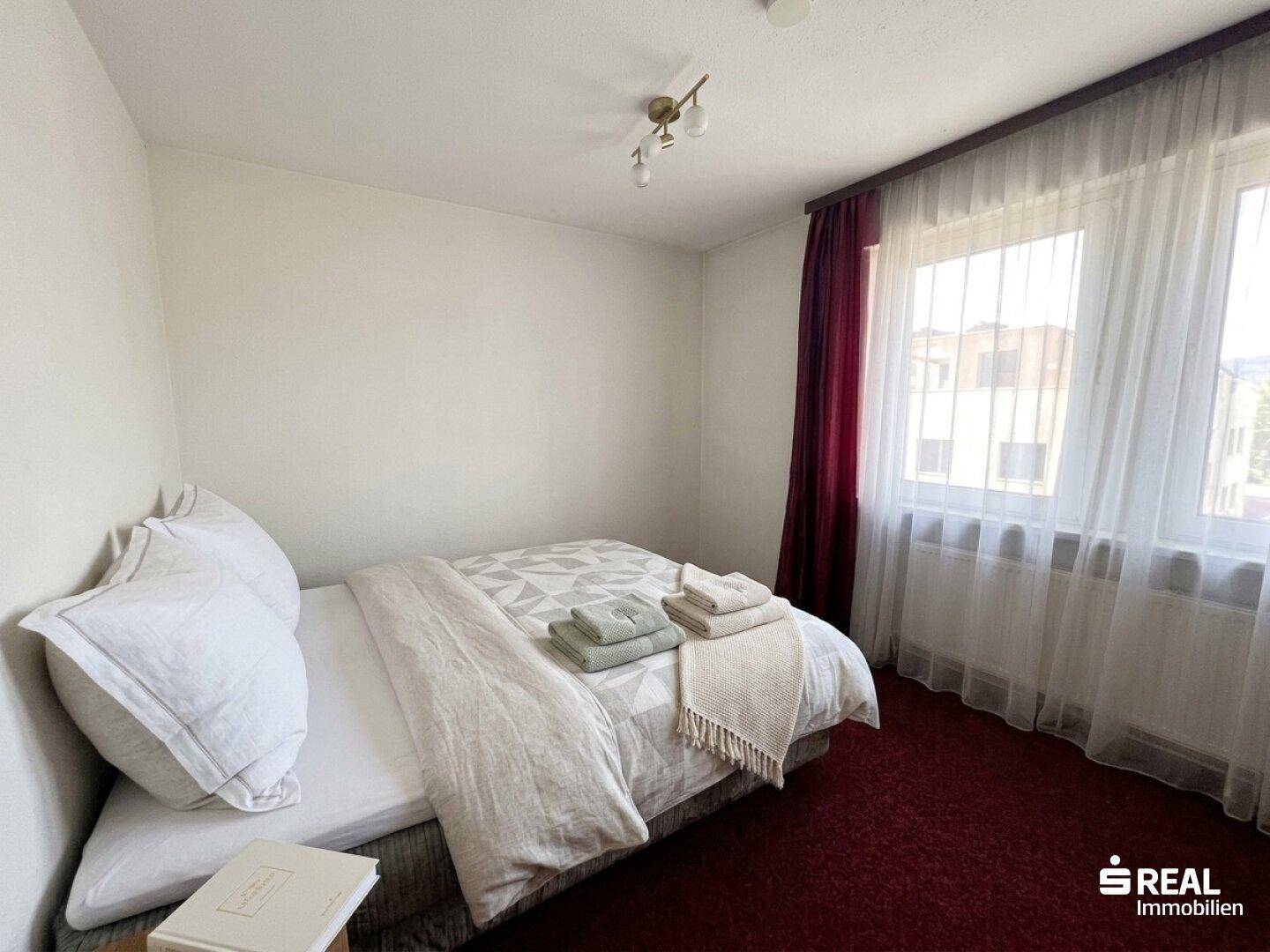 Helles Schlafzimmer mit Einzelbett, rotem Teppichboden und großen Fenstern.