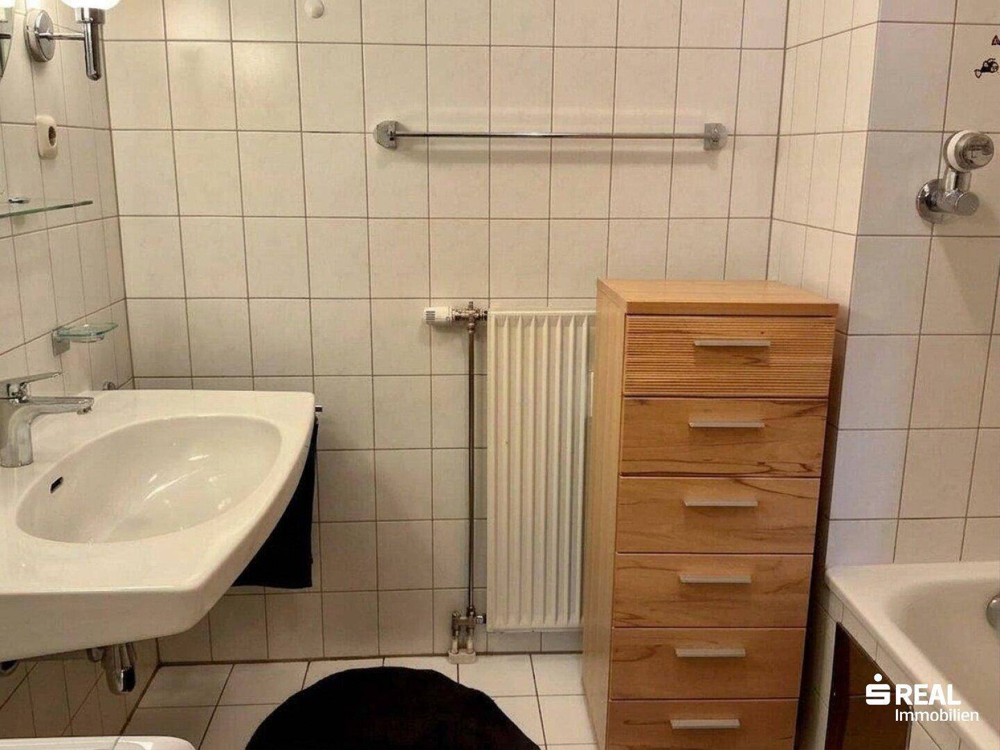 Badezimmer mit weißen Fliesen, Waschbecken, Heizkörper und Holzschrank.