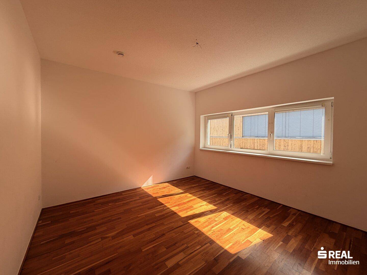 Sonniger Raum mit Holzboden und breitem Fenster, das viel Tageslicht hereinlässt.