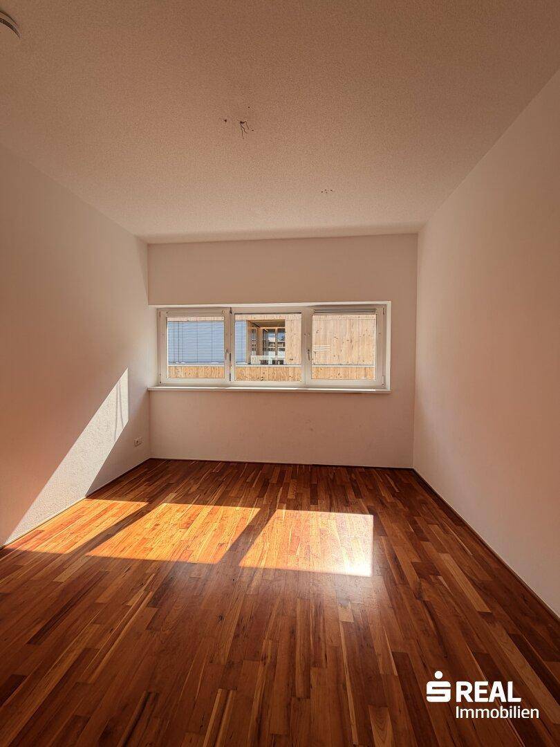 Leeres Zimmer mit Holzboden und einem Fenster, das den Blick nach draußen freigibt.