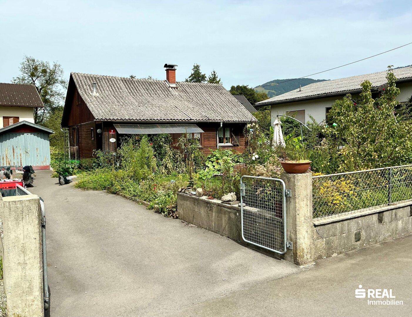 Ländliches Holzhaus mit Garten und Zufahrt, eingebettet in eine grüne Umgebung mit Hügeln.