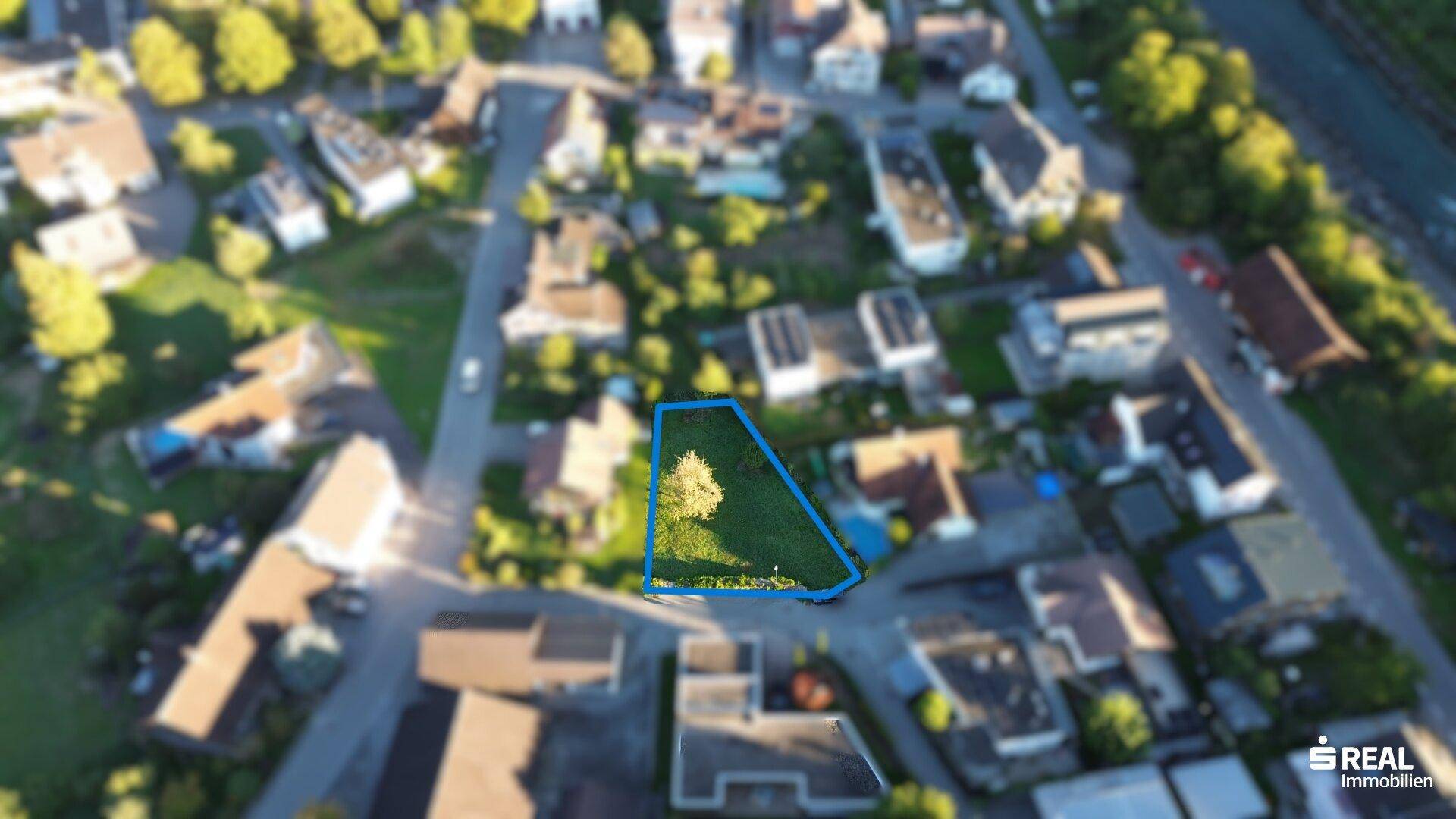 Verschwommene Luftaufnahme eines Grundstücks mit umliegenden Häusern und viel Grün.