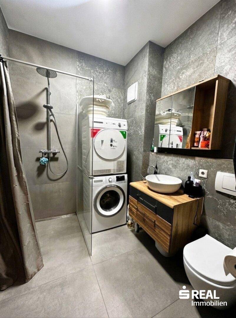 Modernes Badezimmer mit grauen Fliesen, Dusche, Waschbecken und integrierter Waschmaschine.