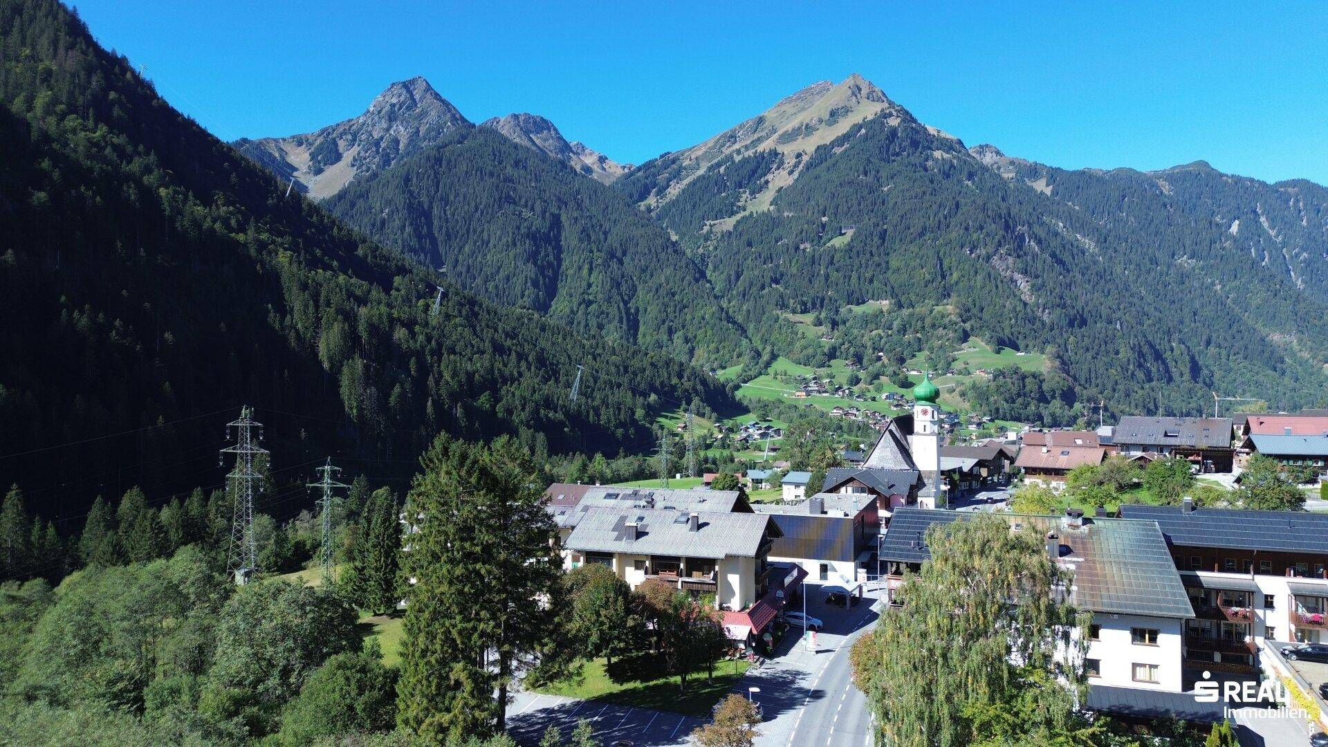 Malerische Luftaufnahme eines Bergdorfes mit Kirche, umgeben von bewaldeten Bergen unter blauem Himmel.
