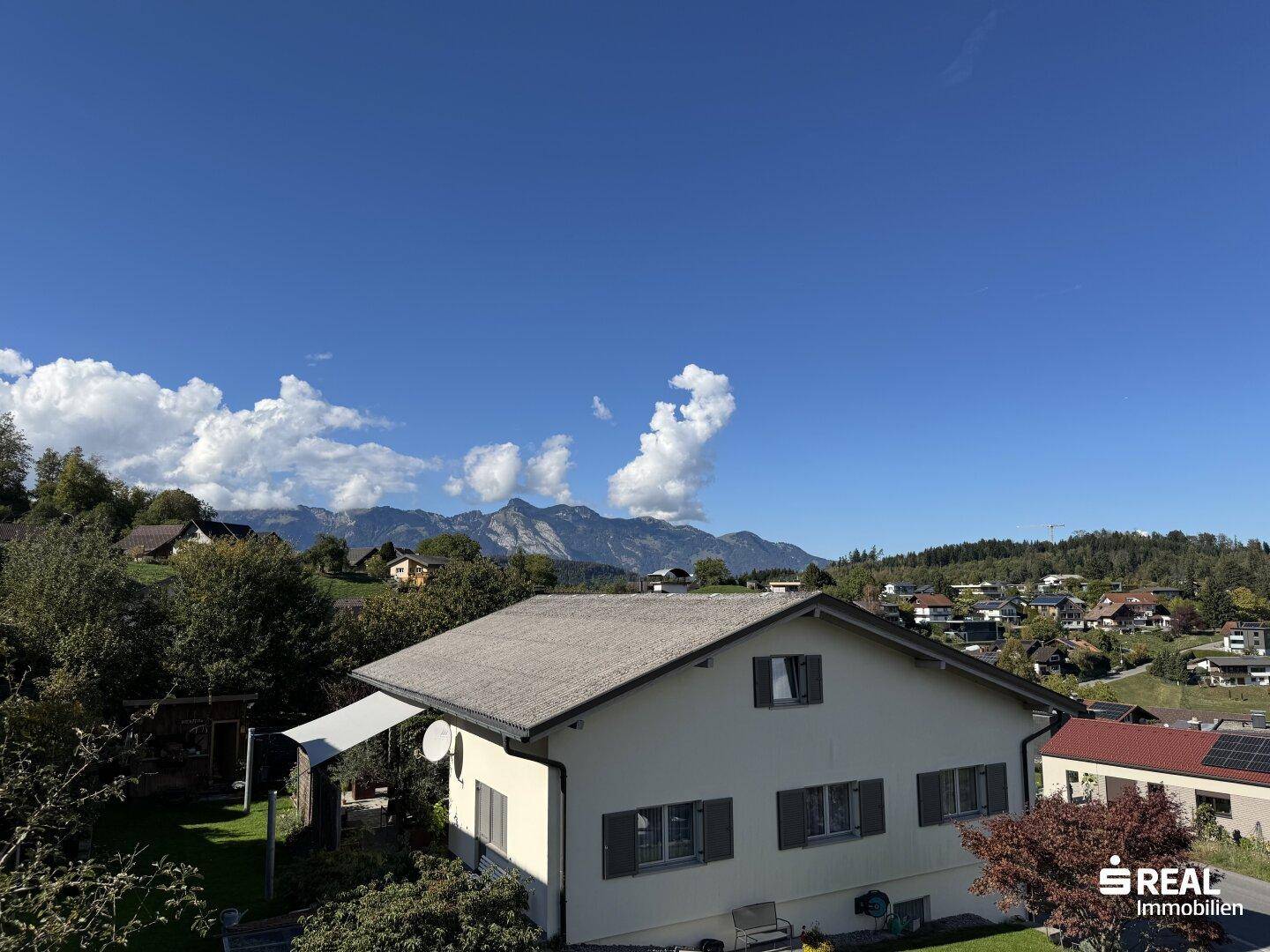 Weitläufiger Panoramablick über eine Siedlung, grüne Hügel und majestätische Berge unter blauem Himmel.