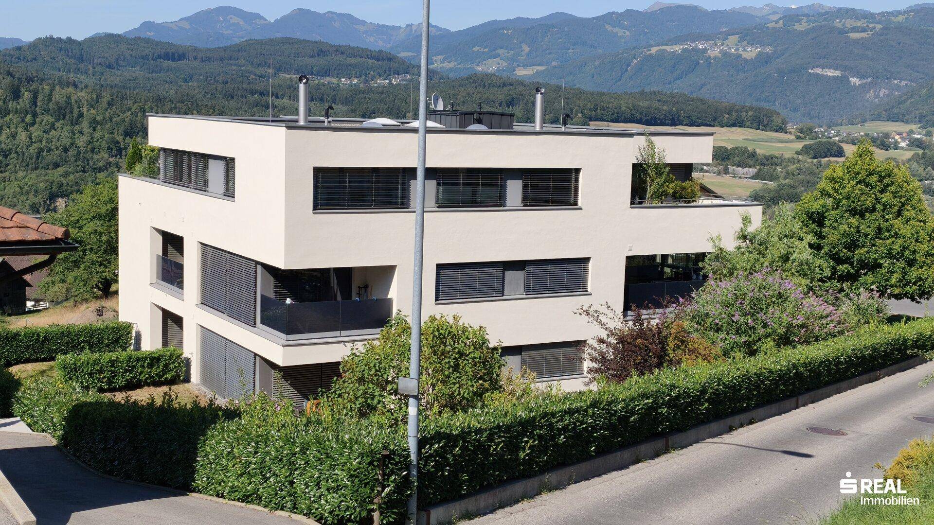 Ansicht des modernen Gebäudes von oben, umgeben von gepflegten Hecken und mit Bergblick.