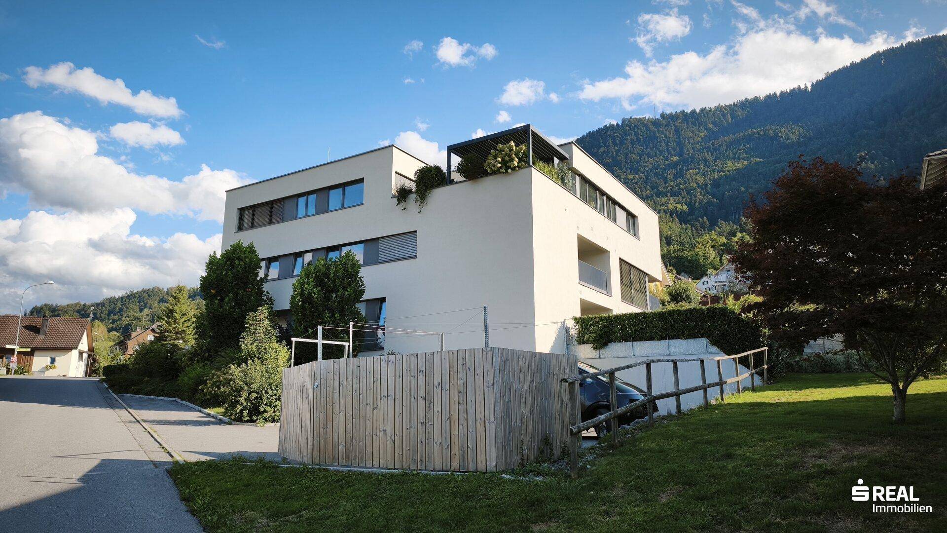 Modernes Wohngebäude mit klaren Linien, Balkonen und Flachdach, eingebettet in eine grüne Umgebung.