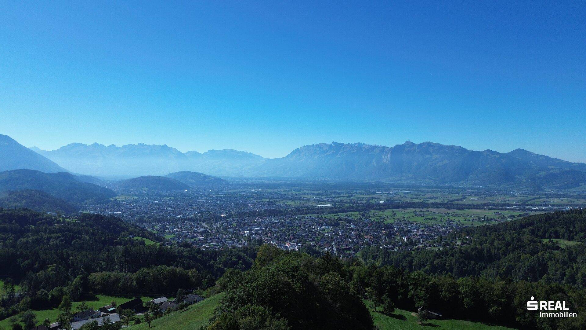 Weitläufige Landschaftsaufnahme mit einer Stadt im Tal und majestätischen Bergen am Horizont unter blauem Himmel.