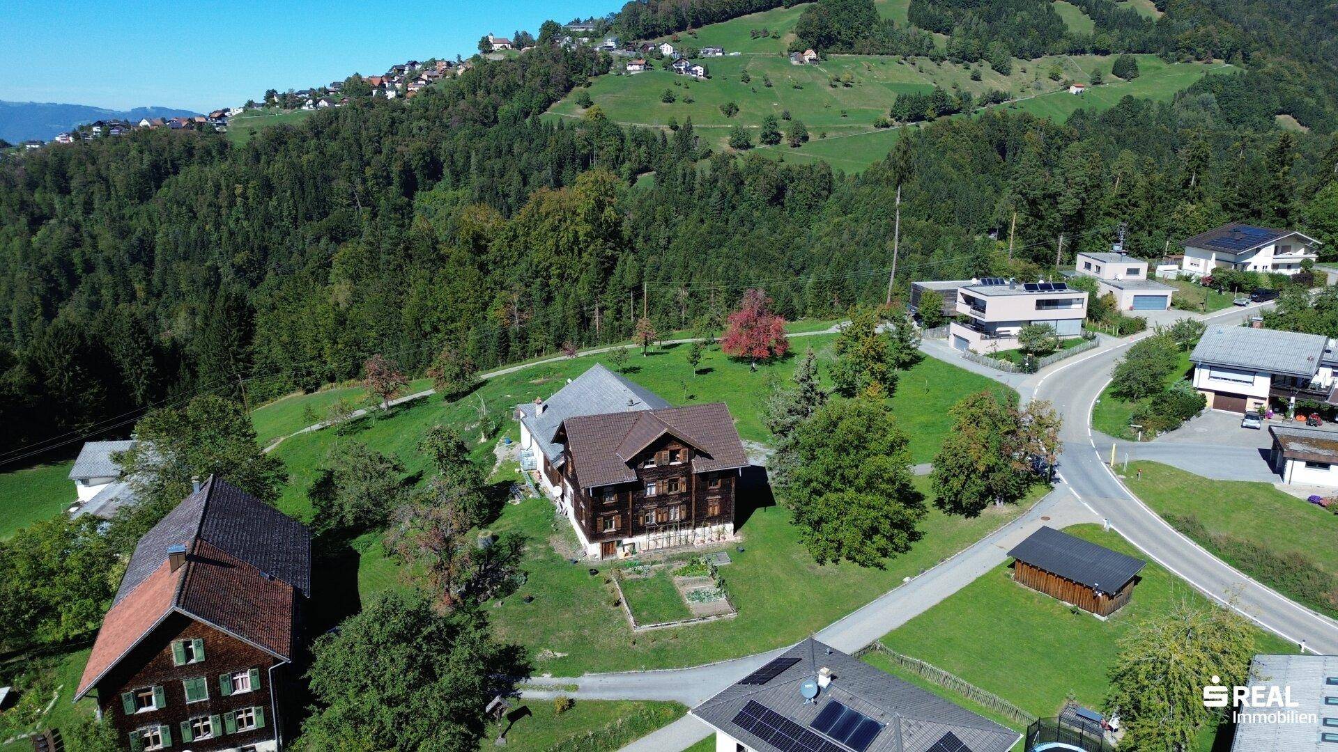 Luftaufnahme zweier traditioneller Häuser mit Gärten, eingebettet in eine grüne Landschaft mit weiteren Gebäuden.