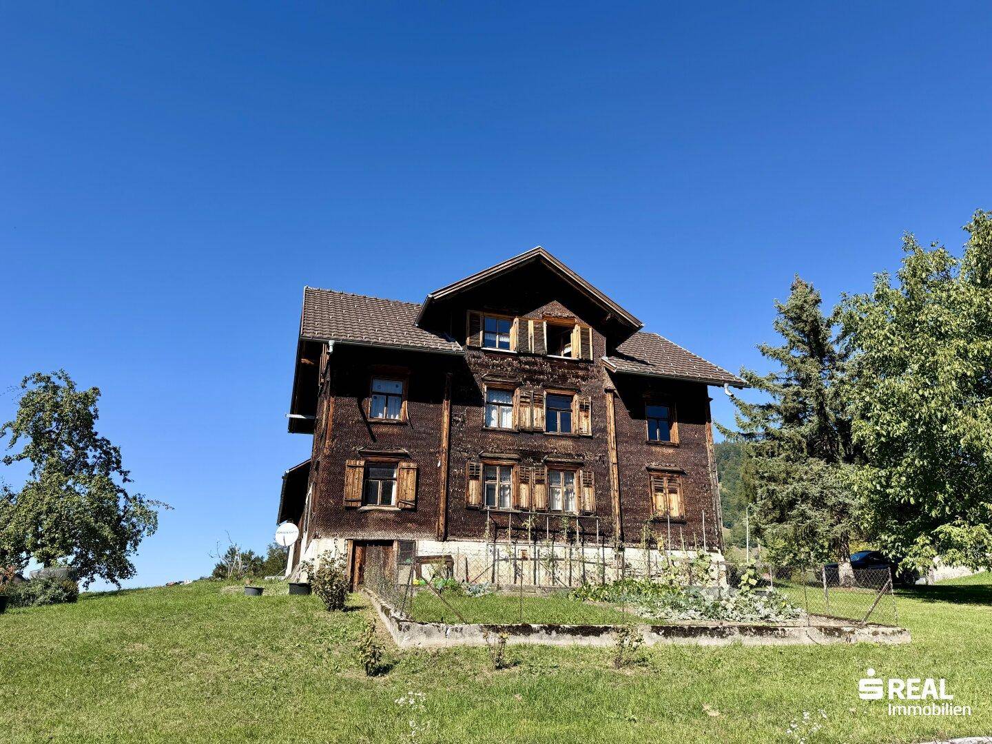 Traditionelles Holzhaus mit Fensterläden und einem kleinen Vorgarten, umgeben von grüner Natur.