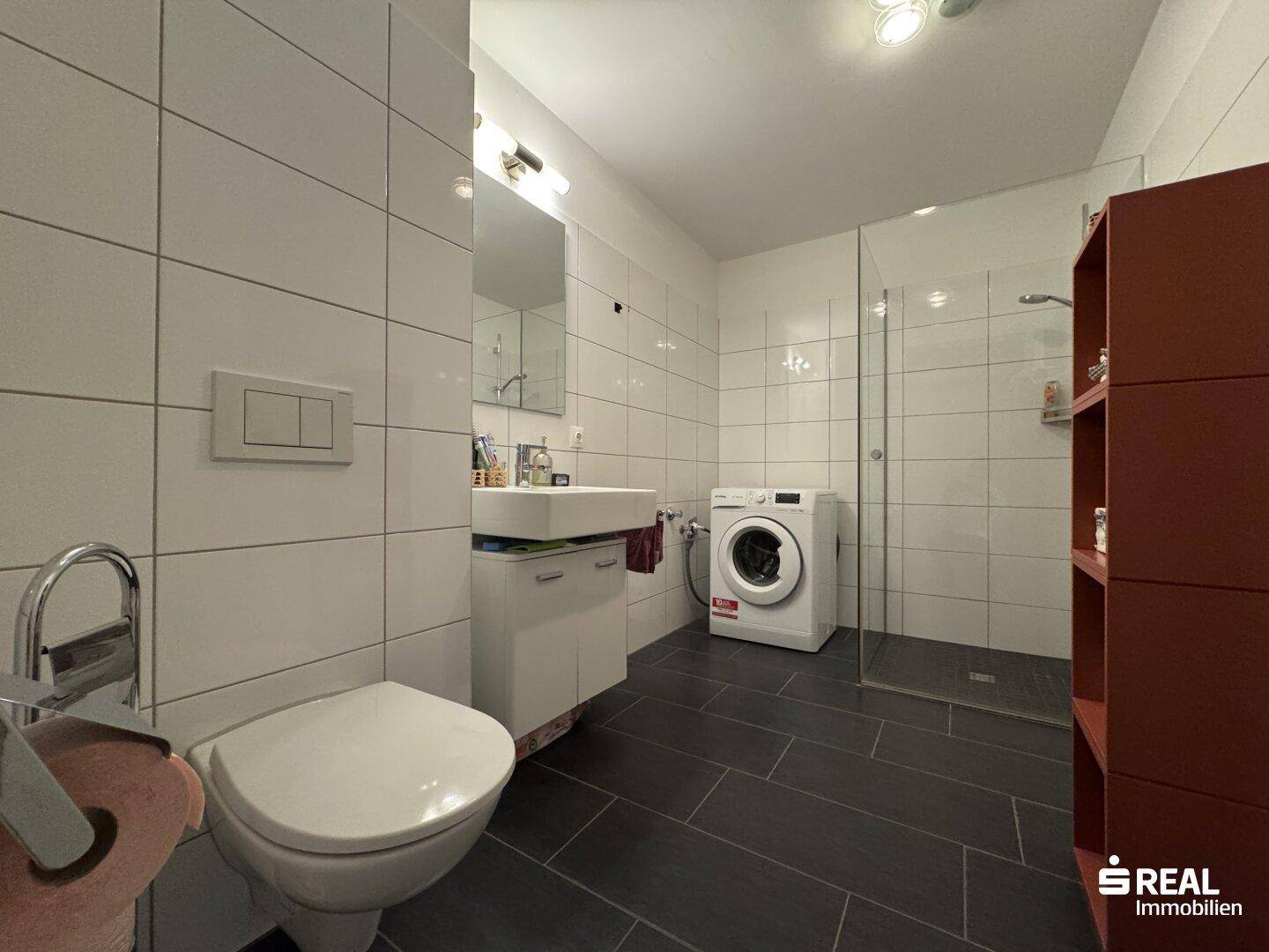 Modernes Badezimmer mit ebenerdiger Dusche, Waschmaschine und dunklen Bodenfliesen.