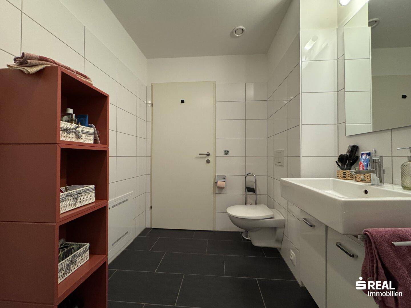 Helles Badezimmer mit weißen Wandfliesen, modernem Waschtisch und praktischem Regal.