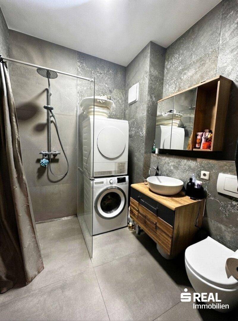 Modernes Badezimmer mit grauen Fliesen, Dusche und Waschmaschine mit Trockner.