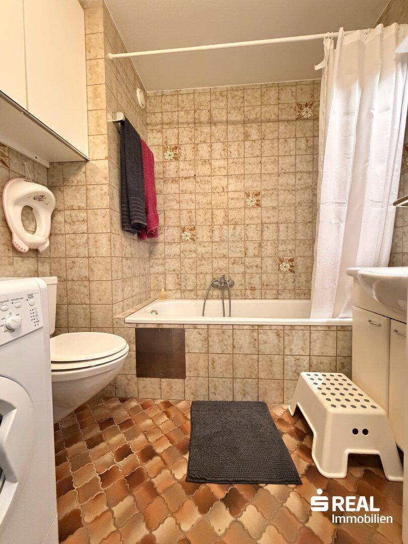 Badezimmer mit Badewanne, WC und Waschmaschine, ausgestattet mit älteren Fliesen.