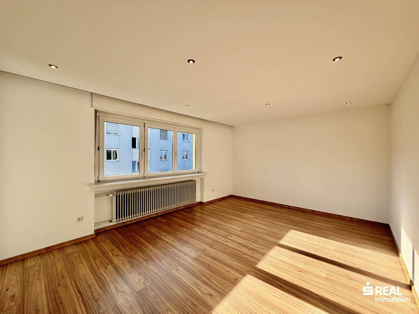 Helles Wohnzimmer mit Holzboden, großen Fenstern und Blick auf die gegenüberliegenden Gebäude.