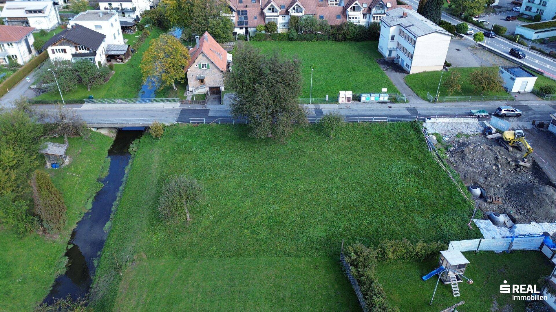 Luftaufnahme eines grünen Baugrundstücks mit roter Umrandung, angrenzendem Bach und umliegenden Häusern.