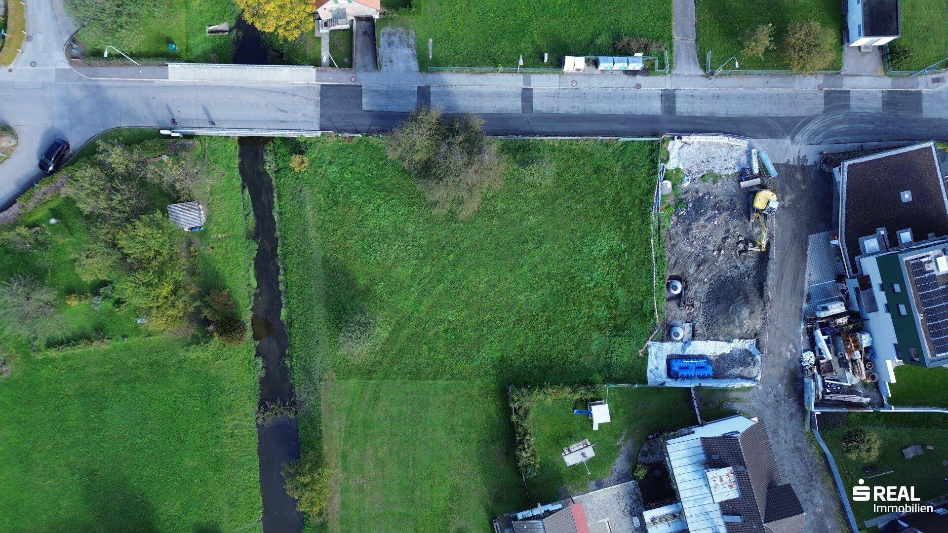 Senkrechte Luftaufnahme des grünen Grundstücks mit angrenzendem Bach, Straße und Baustellenbereich.