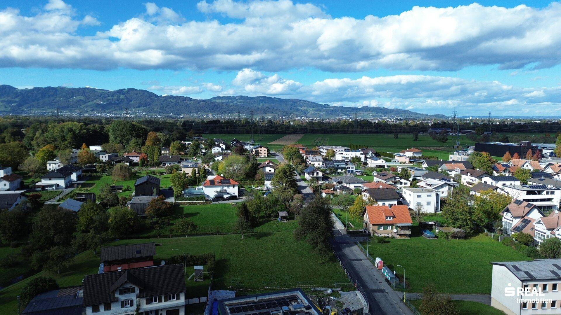 Panoramische Luftaufnahme einer Wohnsiedlung mit weiten Grünflächen und Bergen am Horizont unter blauem Himmel.