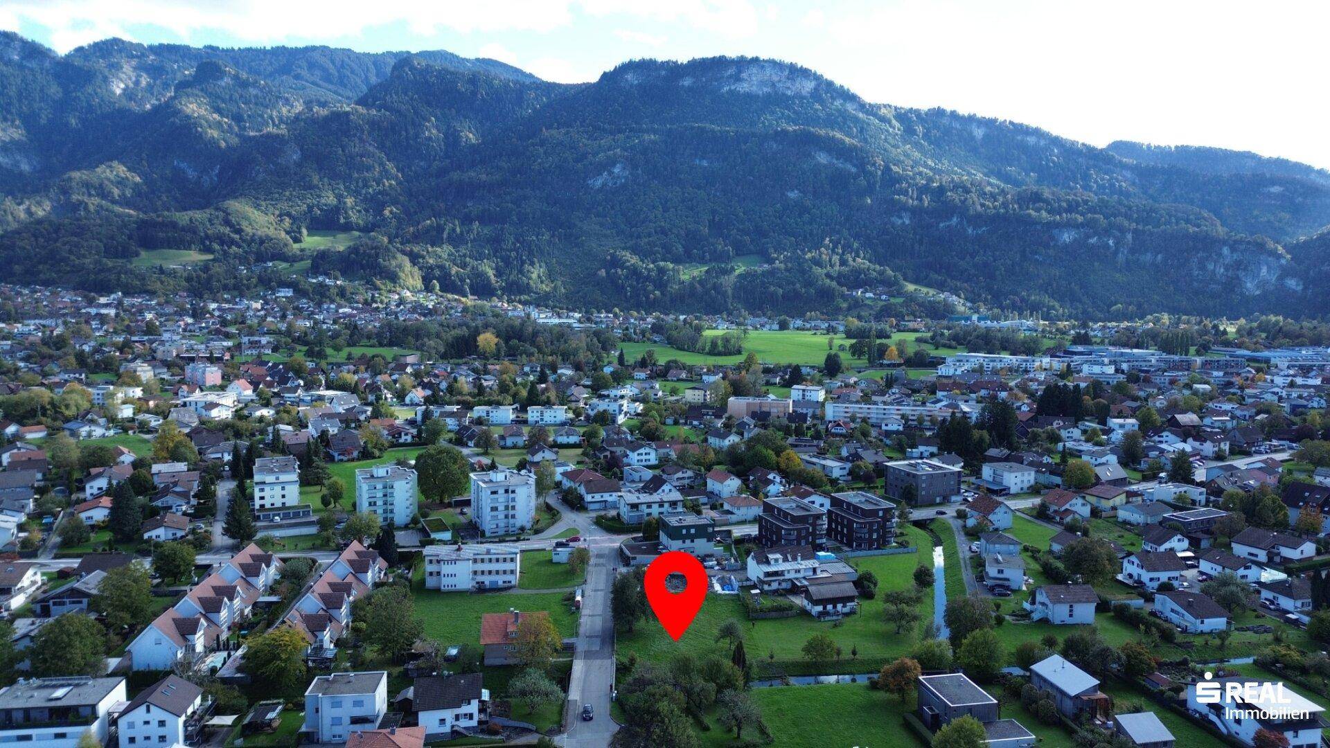 Weitläufige Luftaufnahme der Ortschaft mit markiertem Grundstück und beeindruckender Bergkulisse im Hintergrund.
