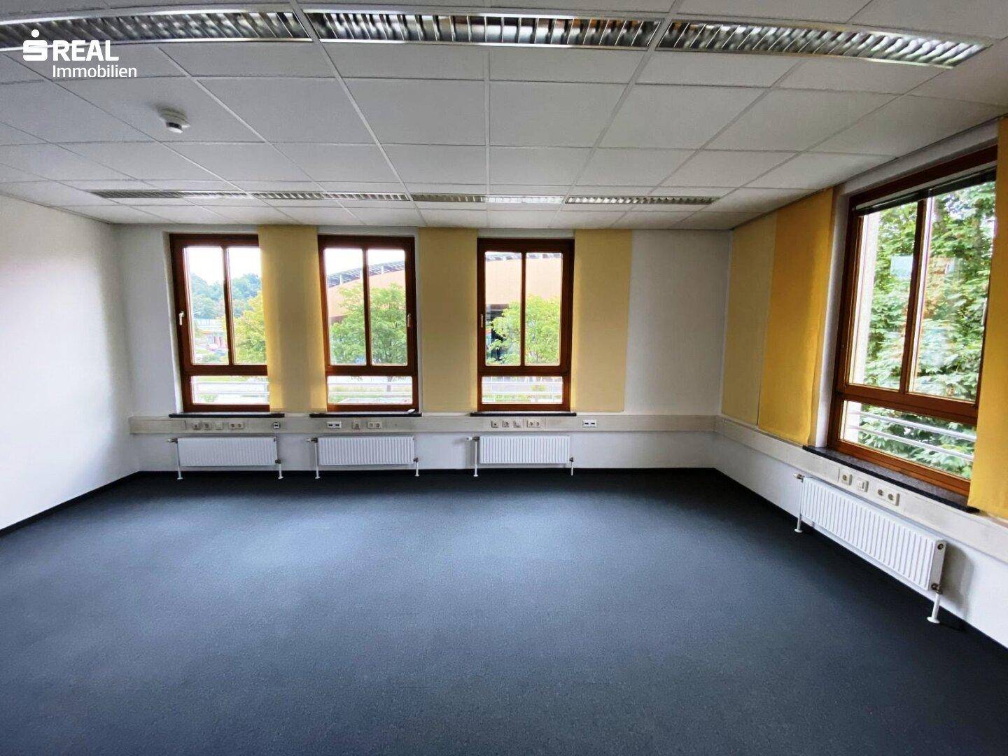 Geräumiges Büro mit Teppichboden, vielen Fenstern und Heizkörpern unter den Brüstungen.