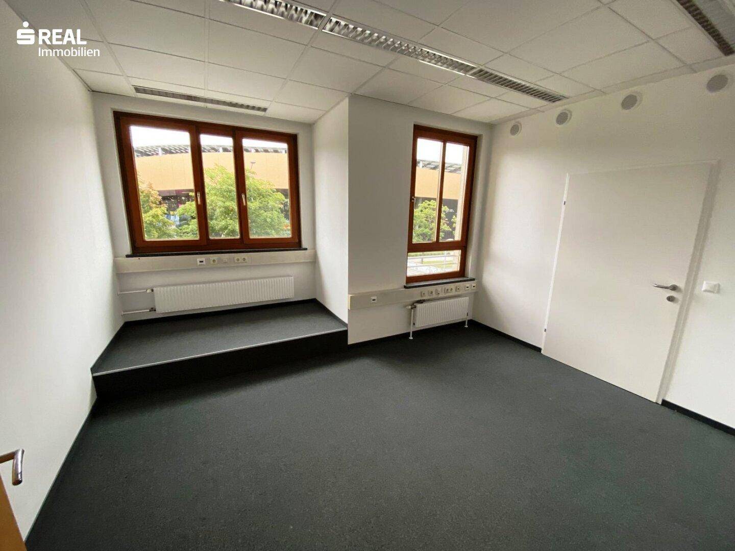 Helles Büro mit Teppichboden, weißen Wänden und zwei Fenstern mit Ausblick.