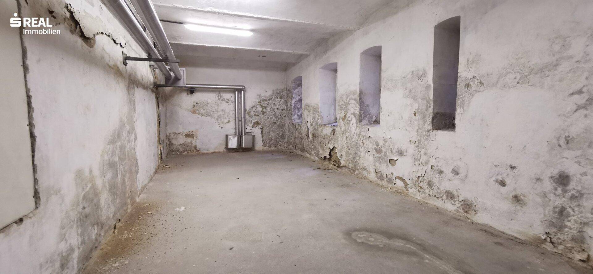 Unsanierter Kellerraum mit feuchten, abblätternden Wänden, Betonboden und sichtbaren Rohrleitungen.