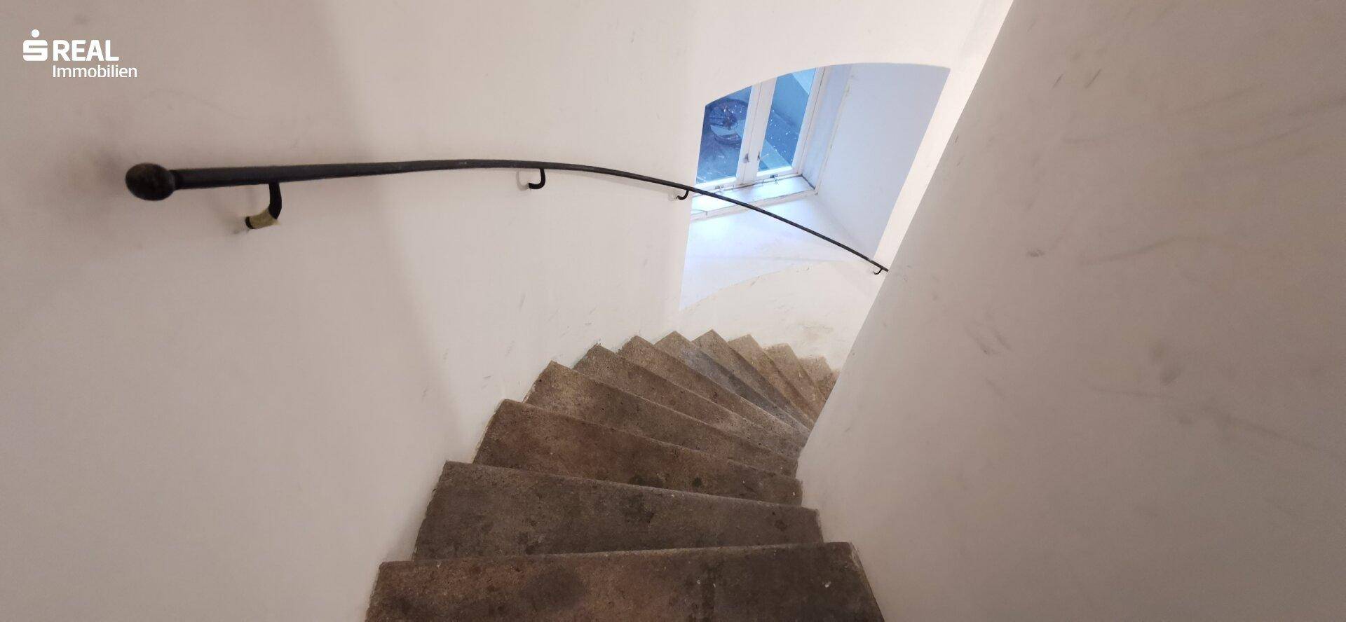 Wendeltreppe mit Steinstufen und einem geschwungenen Handlauf, die zu einem hellen Fenster führt.