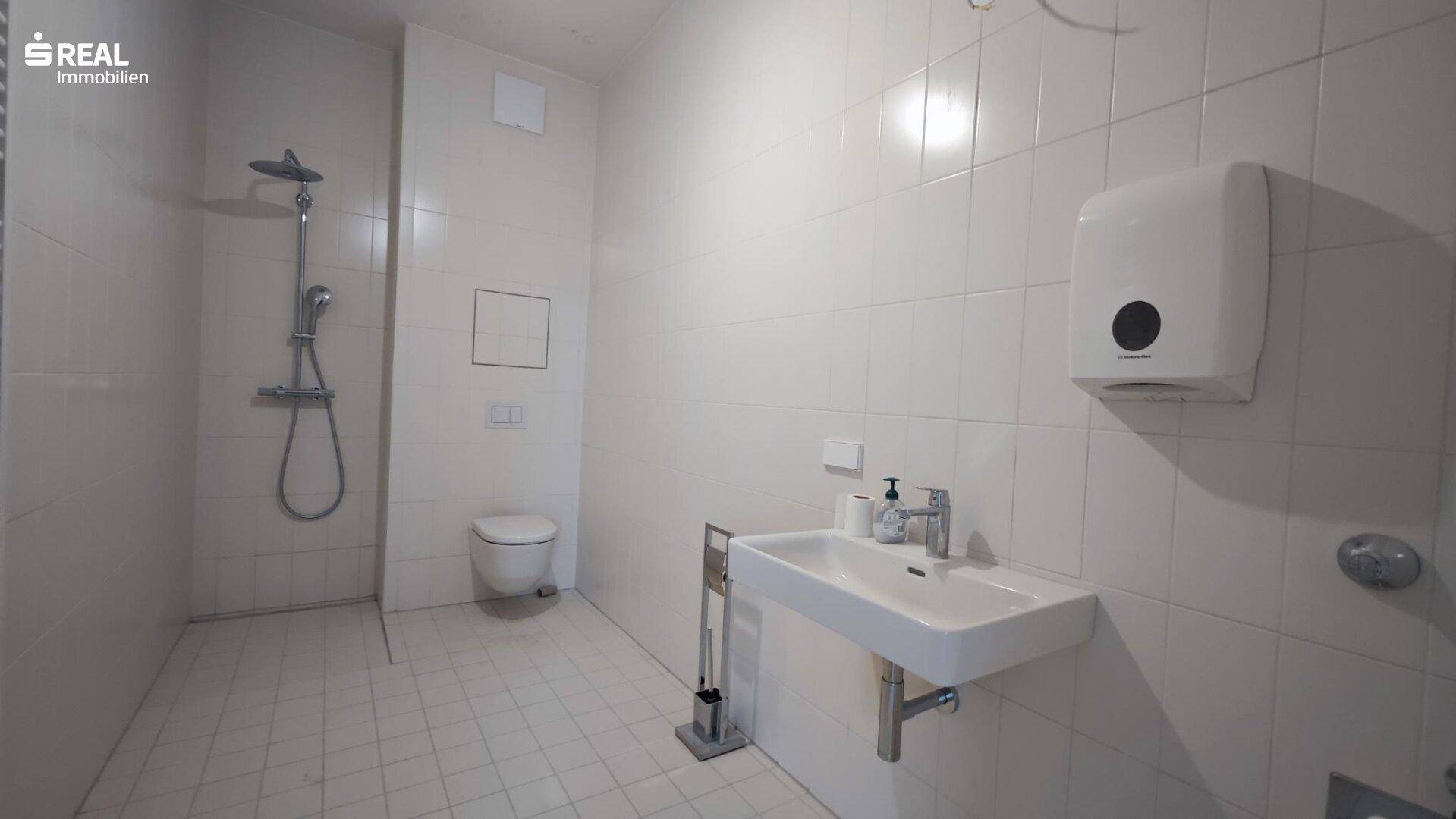 Modernes Badezimmer mit weißen Fliesen, wandhängendem WC, Dusche und Waschbecken, funktional ausgestattet.
