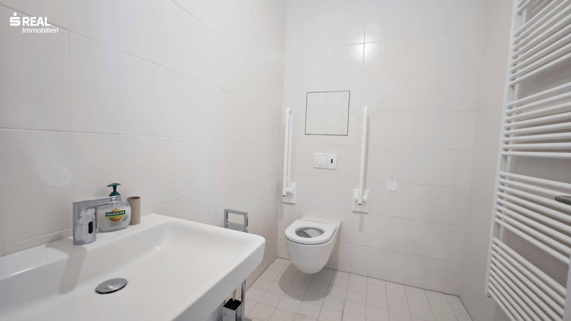 Barrierefreies Badezimmer mit weißen Fliesen, wandhängendem WC, Haltegriffen und einem Handtuchheizkörper.