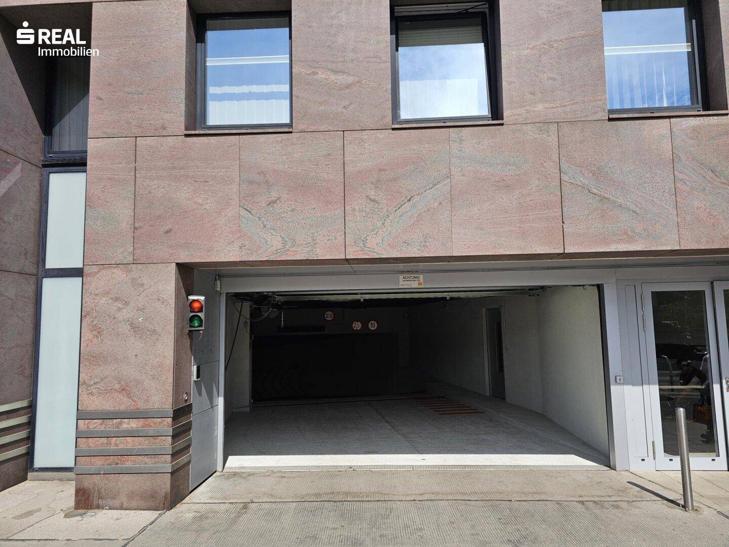 Moderne Gebäudefassade aus Stein mit der Einfahrt zu einer Tiefgarage und großen Fenstern.