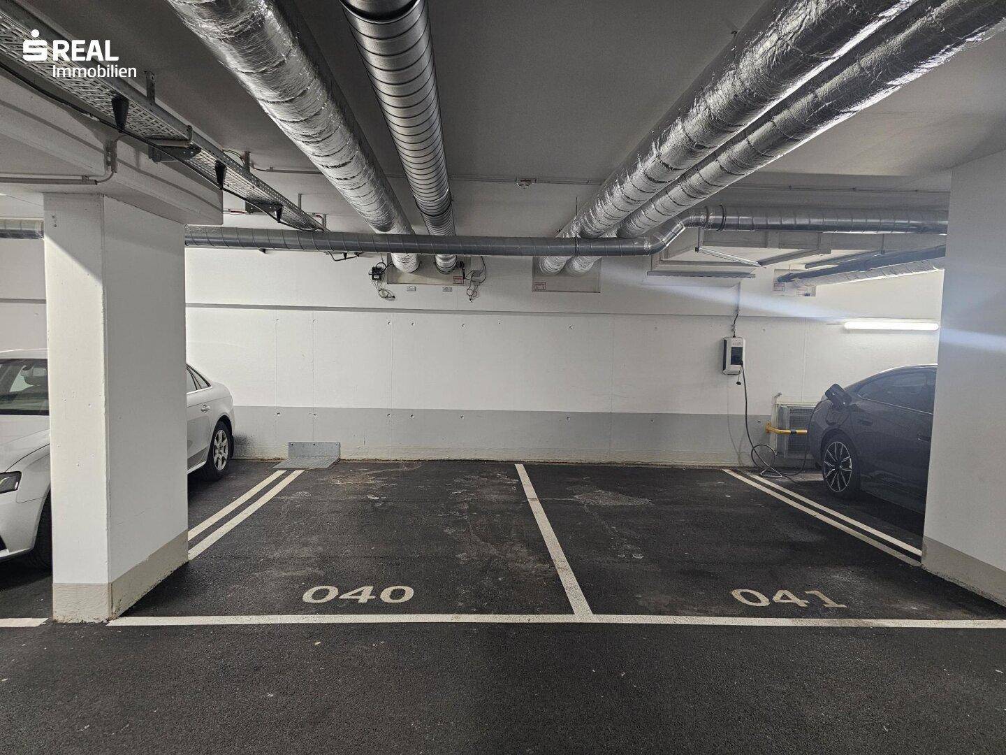 Zwei leere Parkplätze in der Tiefgarage mit einer Ladestation für Elektrofahrzeuge an der Wand.