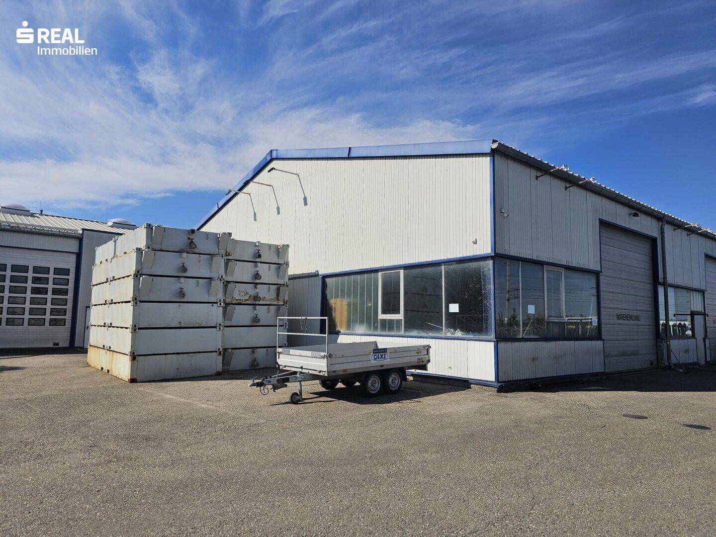 Außenansicht einer großen Lagerhalle mit Rolltoren und gestapelten Containern auf dem Vorplatz.