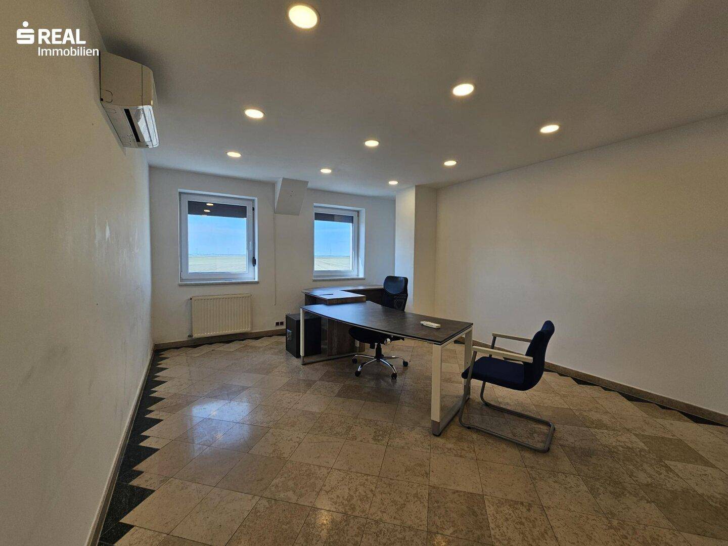 Möbliertes Büro mit Schreibtisch, Bürostühlen und Fenstern mit Blick nach draußen.