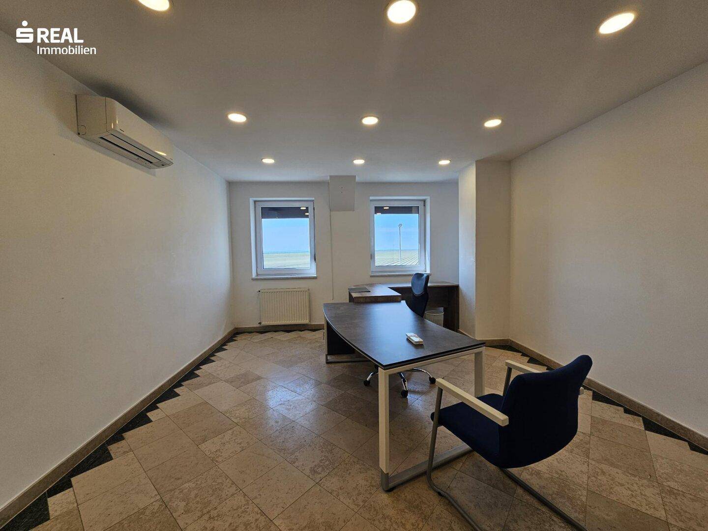 Möbliertes Büro mit Schreibtisch, Bürostühlen und Fenstern mit Blick nach draußen.