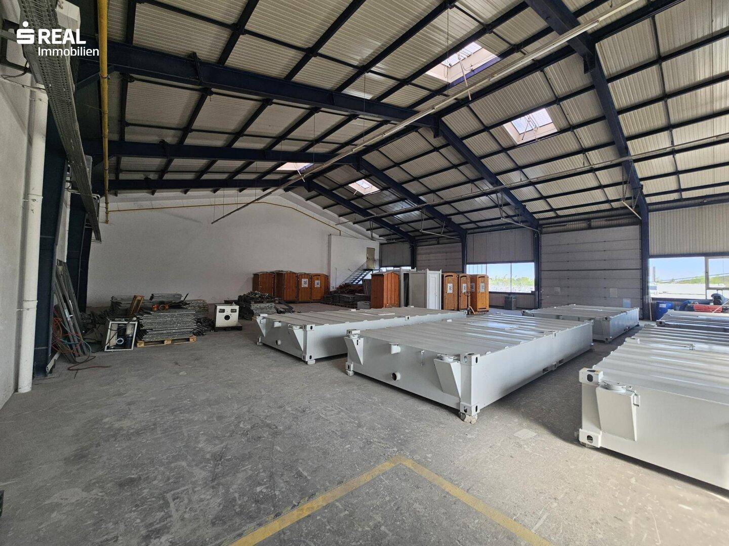 Weitläufige Lagerhalle mit hohen Decken, Oberlichtern und gestapelten Containern.