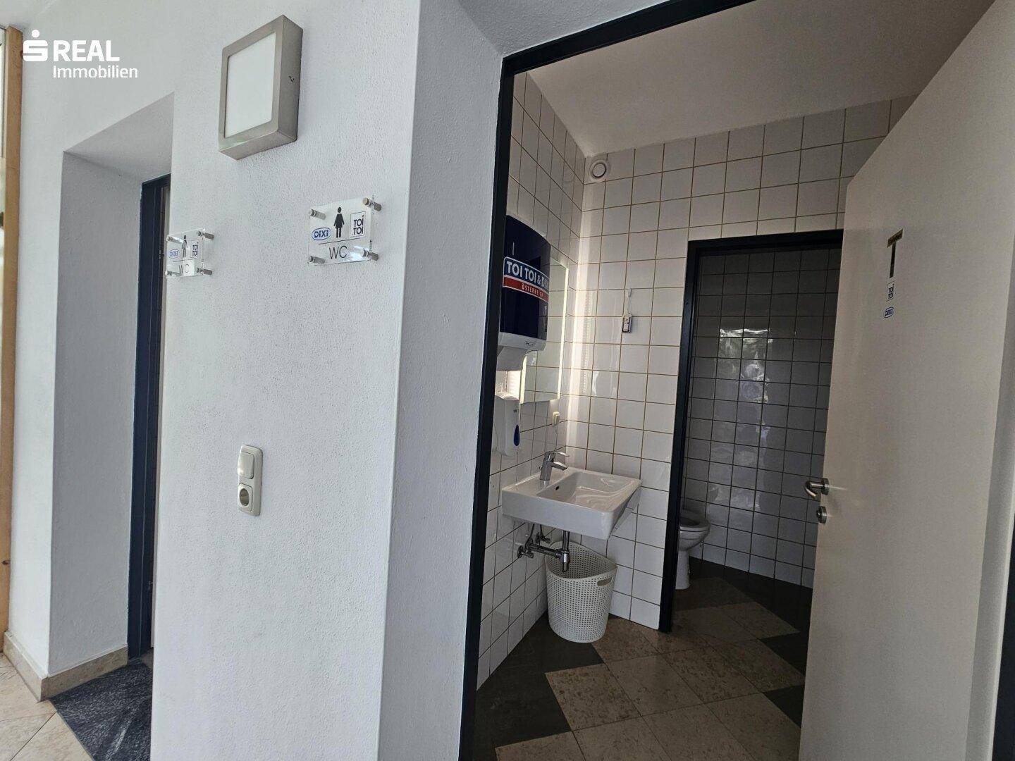 Einfacher Toilettenbereich mit Waschbecken, Spiegel und weißen Wandfliesen.