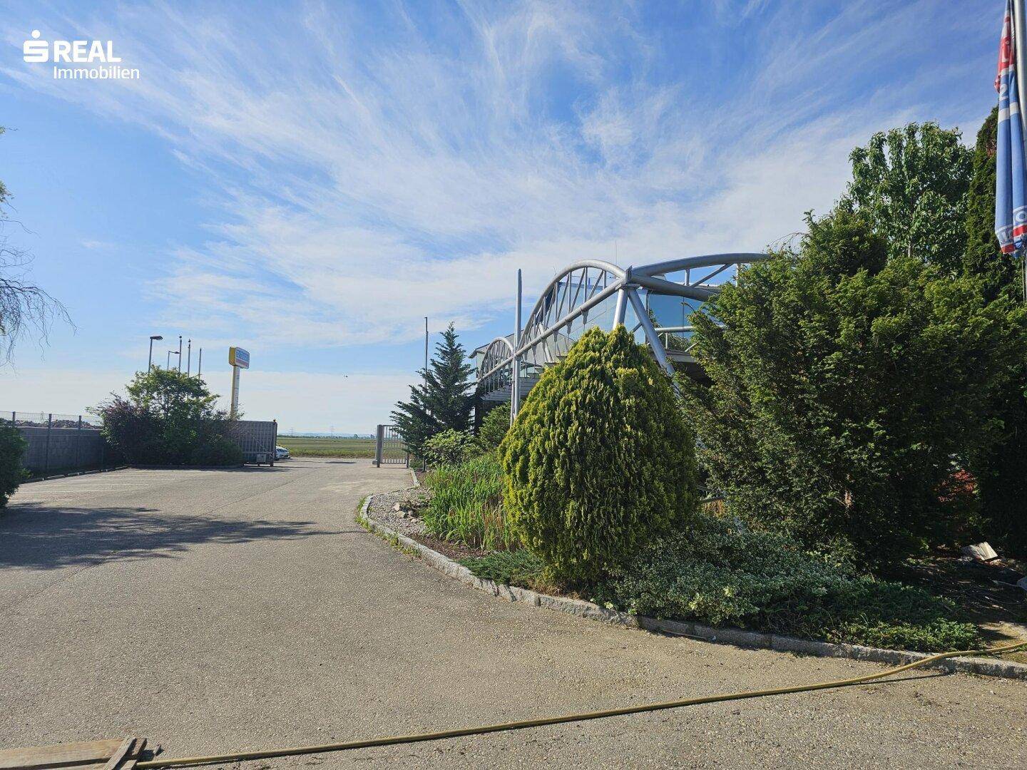 Zufahrt zum Gebäude mit gepflegter Bepflanzung und einem modernen Glasbau im Hintergrund.