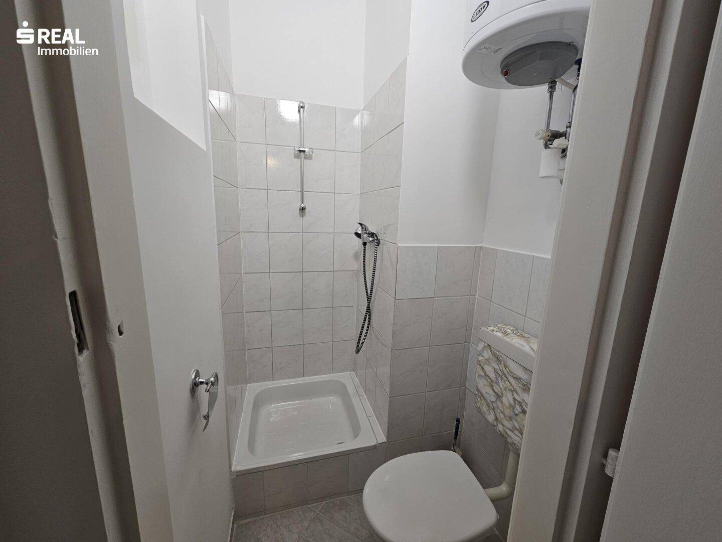 Kleines Badezimmer mit Duschkabine, Toilette und einem Warmwasserboiler an der Wand.