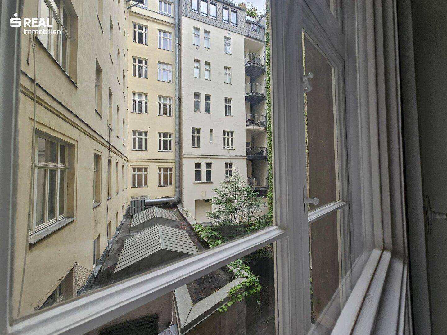Blick aus dem Fenster auf einen ruhigen Innenhof mit Bäumen und umliegenden Gebäuden.