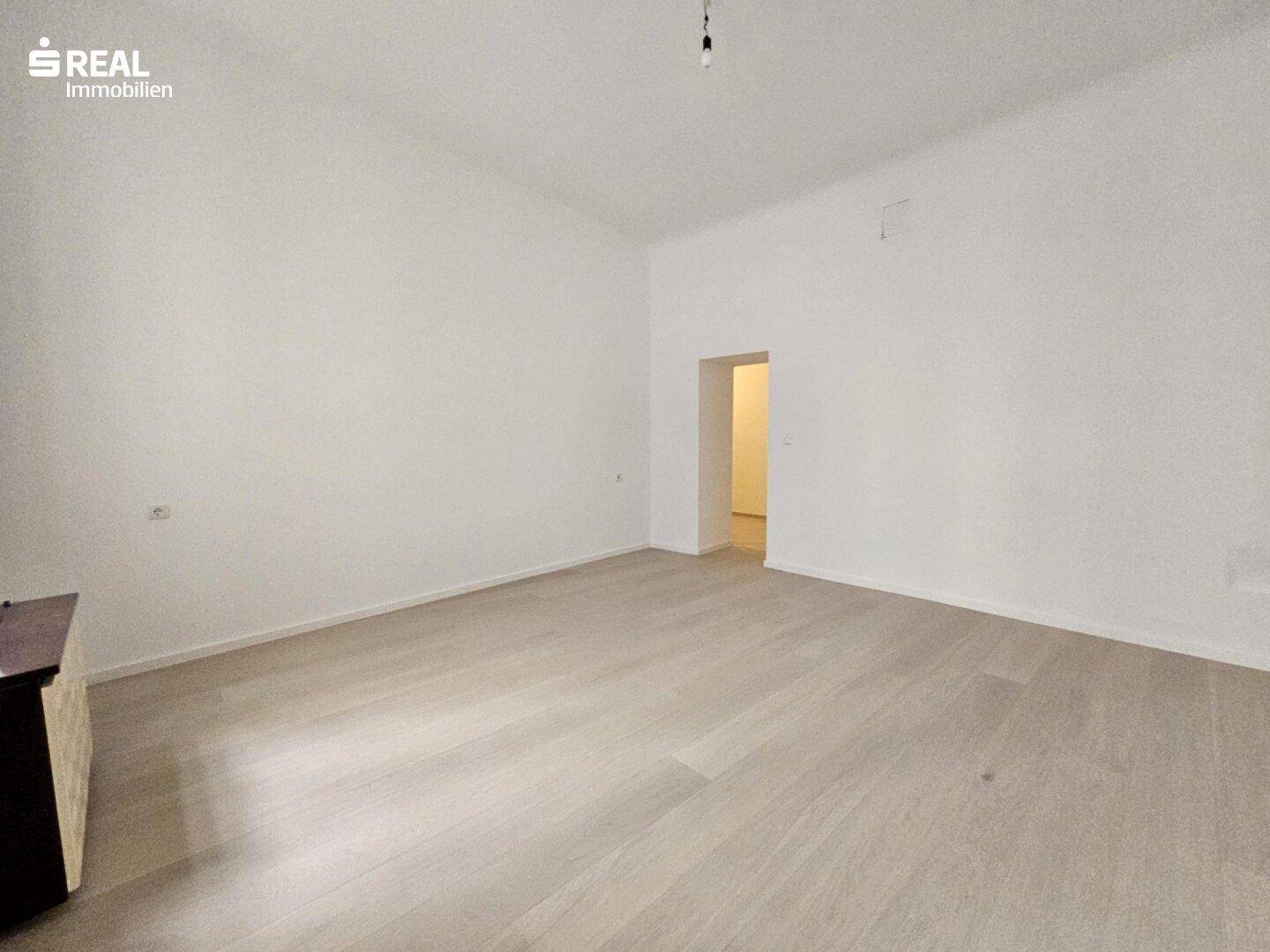 Geräumiger, heller Raum mit Holzboden und einem Durchgang zu einem weiteren Bereich.