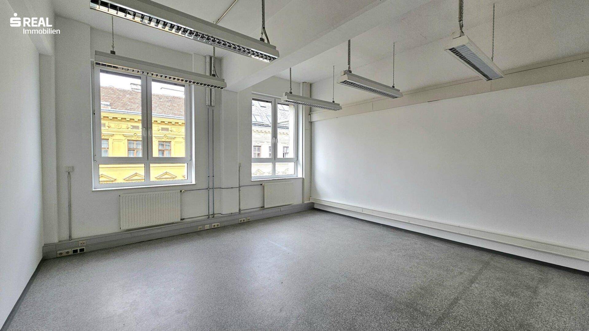 Leerer Büroraum mit Betonboden, weißen Wänden und großen Fenstern mit Blick nach draußen.