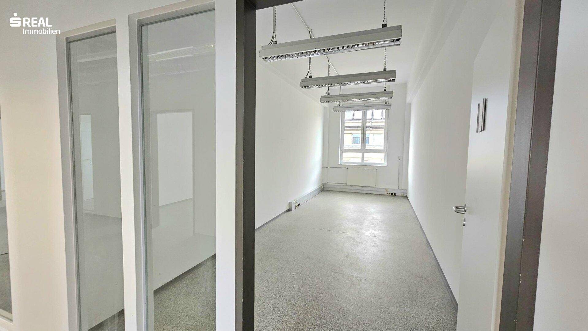 Langer Büroraum mit Betonboden, weißen Wänden und mehreren Fenstern, die Tageslicht spenden.