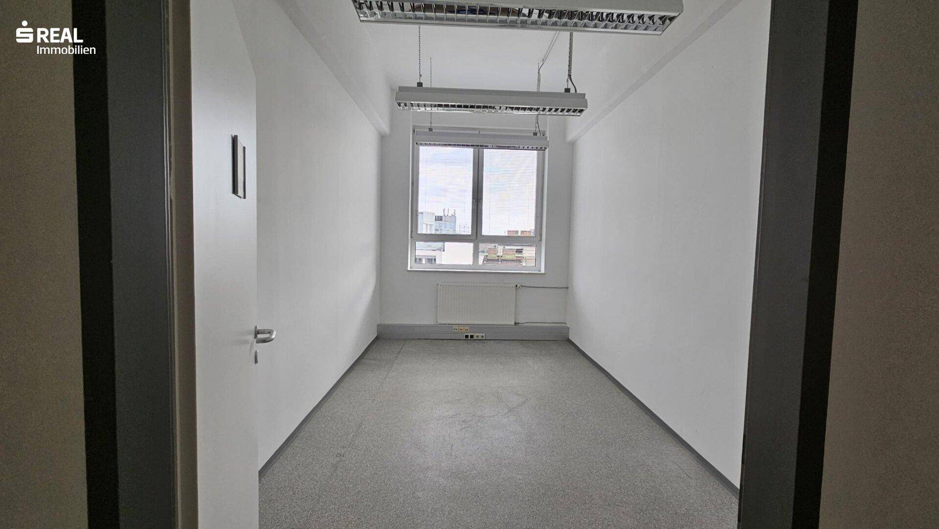 Kleiner Büroraum mit einem Fenster, das viel Tageslicht hereinlässt, und einem Betonboden.