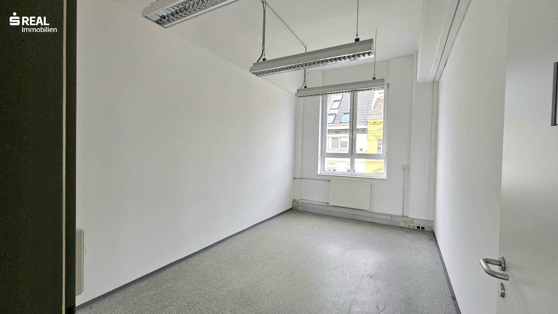 Leerer Büroraum mit Betonboden, weißen Wänden und einem Fenster mit Blick auf Dächer.