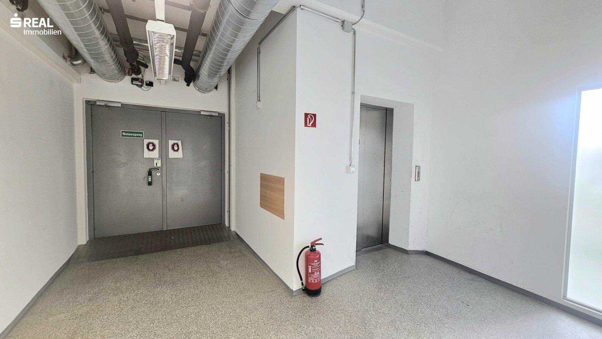 Flur mit Aufzug, Brandschutztüren und einem Feuerlöscher, typisch für ein Bürogebäude.